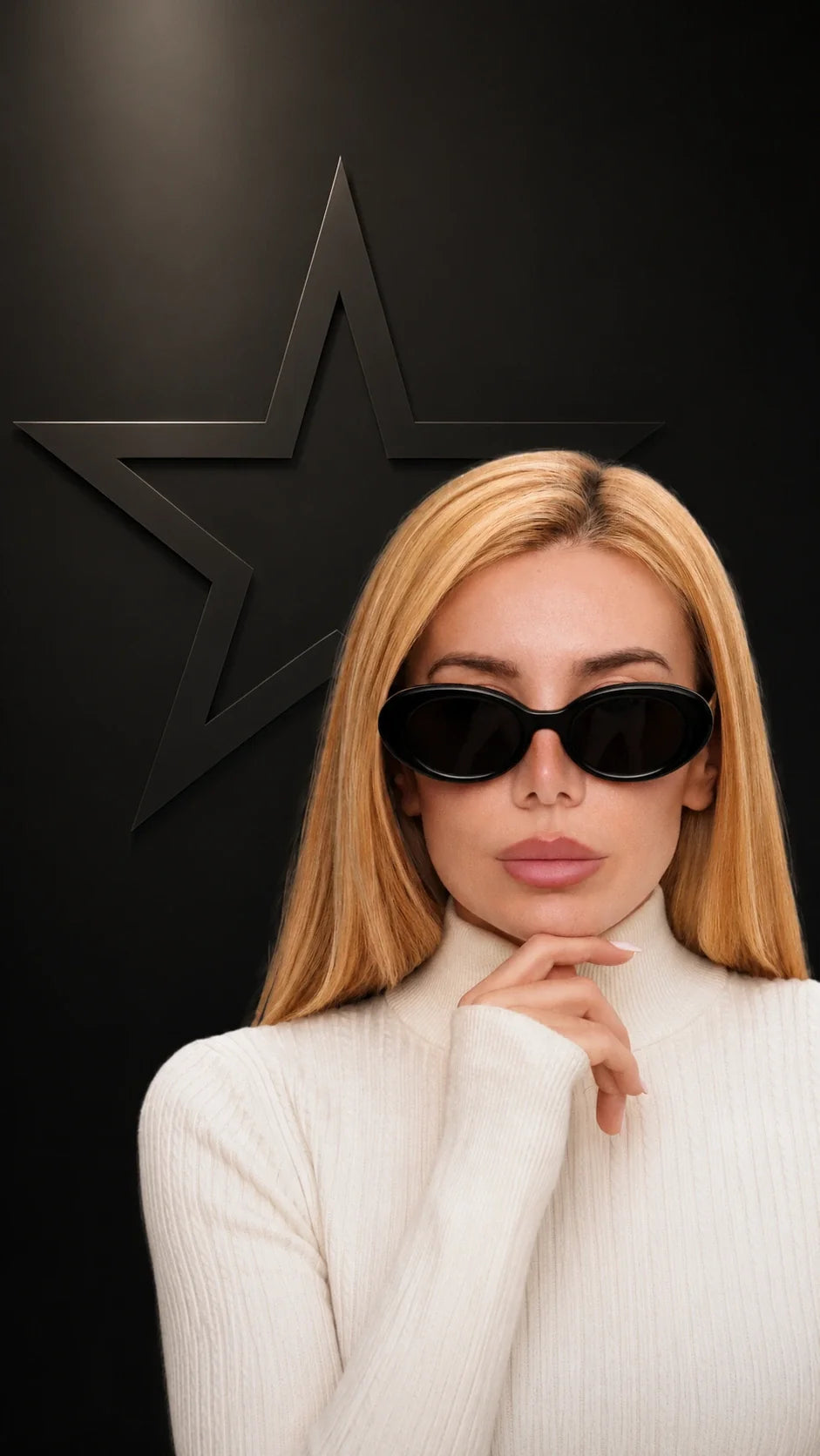 A woman with blonde hair wearing black cat-eye sunglasses and a white ribbed turtleneck sweater, posing against a dark background with a large black star emblem.