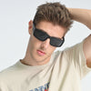 Man wearing black sunglasses and a beige t-shirt against a light gray background
