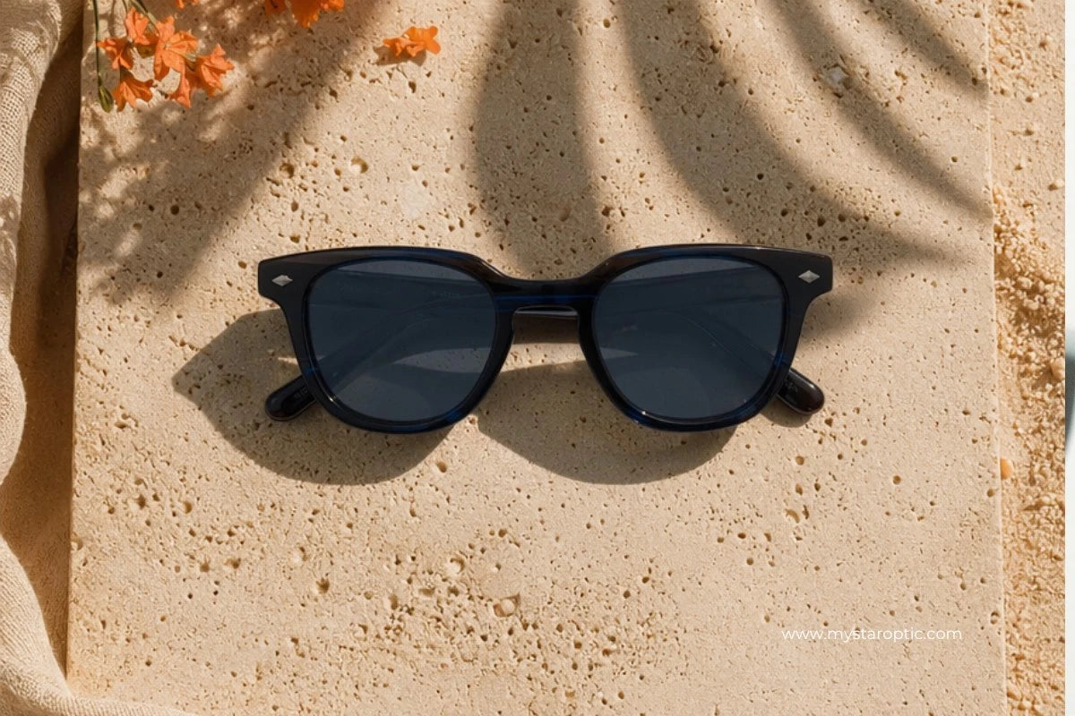 Black square-frame sunglasses with dark lenses resting on a textured beige stone surface with warm sunlight and leaf shadows.