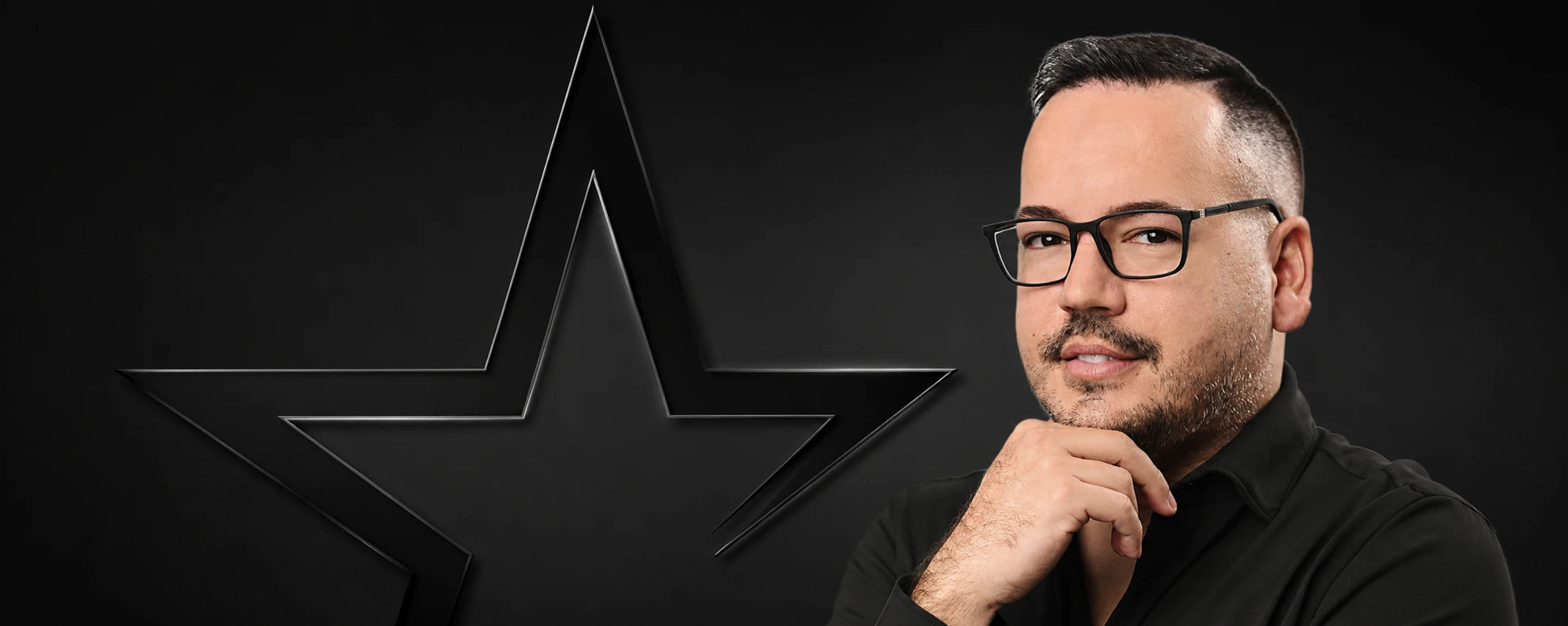 A man with glasses and a beard, wearing a black shirt, posing next to a large black star logo on a dark background.